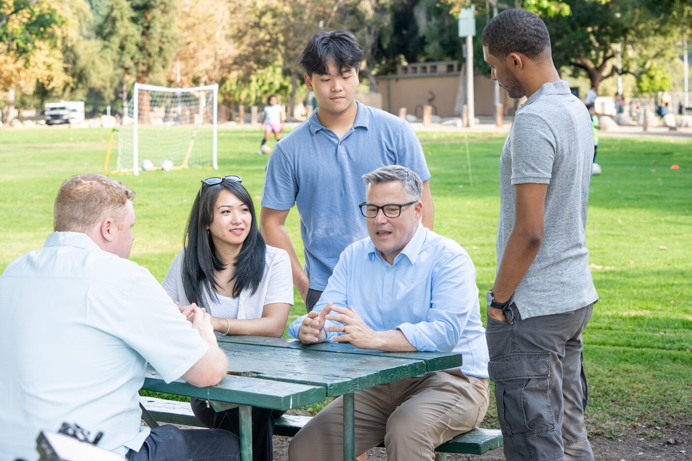 Geoff Baum seated at an outdoor picnic table in conversation with a group of PCC students.